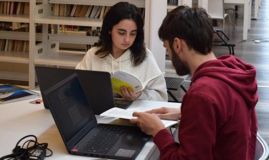 Fotografía en la cual se ven dos estudiantes de la FIC trabajando en la biblioteca, una de ellas con una revista académica sobre bibliotecología.