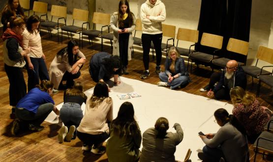 Fotografía. En un salón, un grupo de personas trabajan en torno a un papelógrafo apoyado en el piso. Algunas están paradas, otras sentadas en el suelo y otras de rodillas. Algunas tienen marcadores y escriben en el papel. 