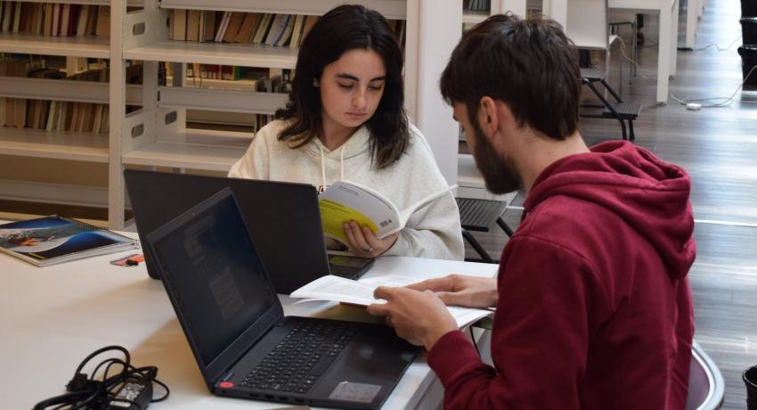 Fotografía en la cual se ven dos estudiantes de la FIC trabajando en la biblioteca, una de ellas con una revista académica sobre bibliotecología.